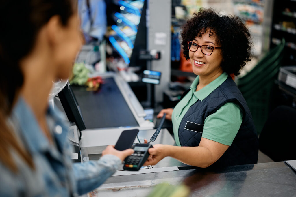 A smiling retail worker wearing glasses and a store uniform processes a contactless payment at a checkout counter, holding a payment terminal toward a customer's phone, with store shelves visible in the background.
