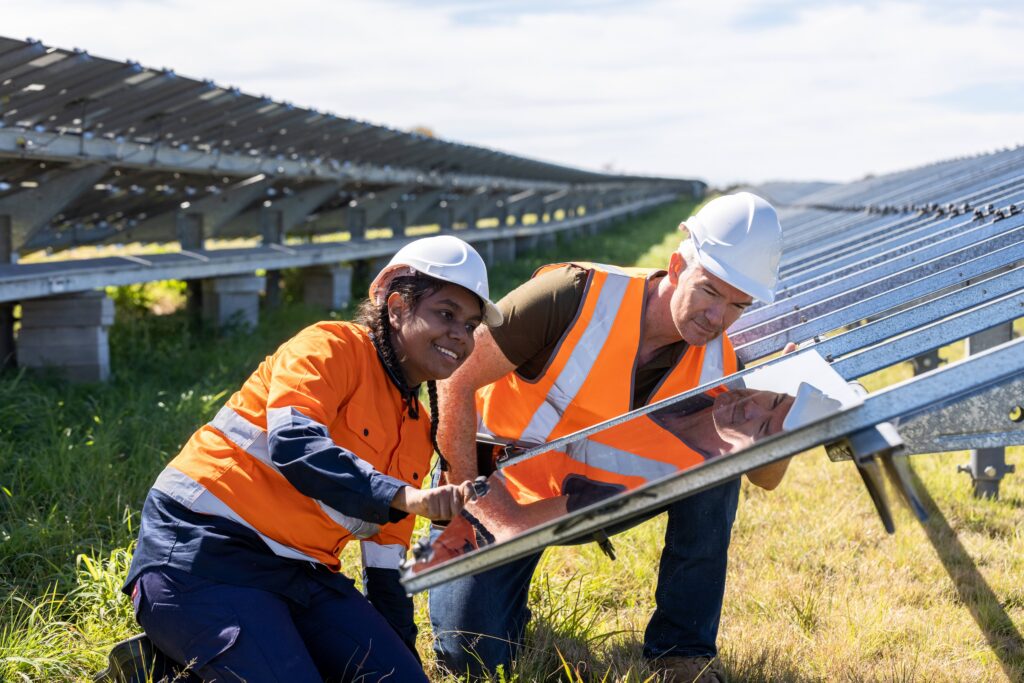 Two workers wearing hi-vis vests and hard hats crouch together while installing a solar panel, with rows of solar panel arrays extending into the distance across a grassy field.