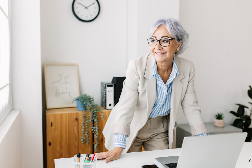 A person with gray hair and glasses stands at a desk in a bright office, smiling and looking toward a window, with a laptop open in front of them.
