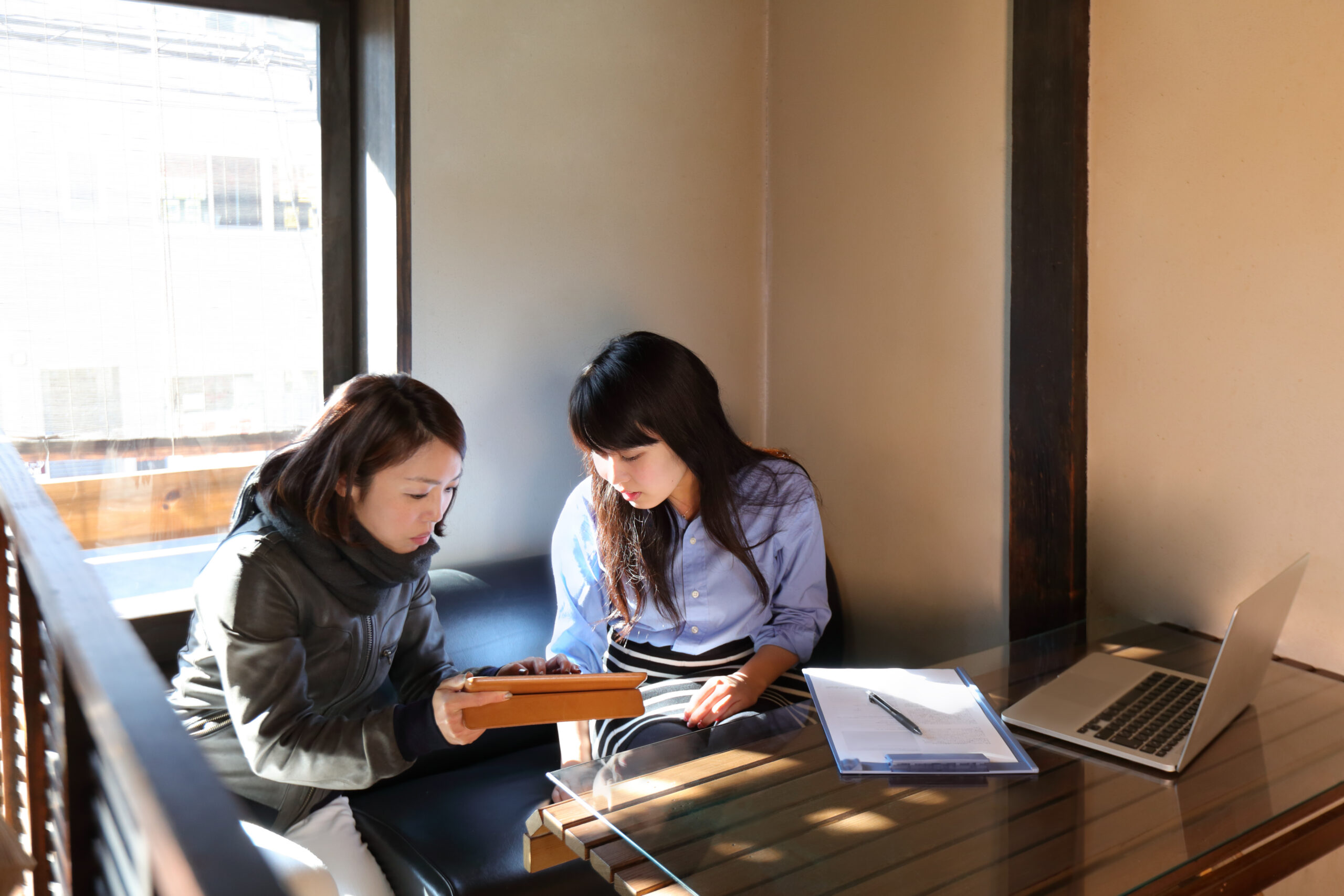 窓際のソファに座る2人が、1人がタブレットを持ちながら並んで画面を見ている様子。テーブルの上にはラップトップと、紙とペンが挟まれたクリップボードが置かれている。