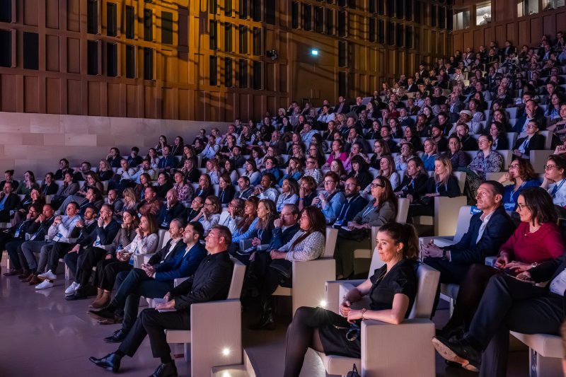 Un grand auditorium rempli de gens enthousiastes attendant que l'orateur prenne la parole sur scène lors d'un grand événement.