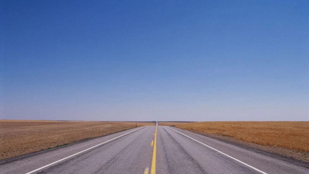 Eine lange, gerade Asphaltstraße mit einer durchgezogenen gelben Linie in der Mitte und weißen Seitenstreifen, die sich bis zum Horizont erstreckt. Links und rechts der Straße erstrecken sich weite, trockene Felder unter einem klaren, blauen Himmel.
