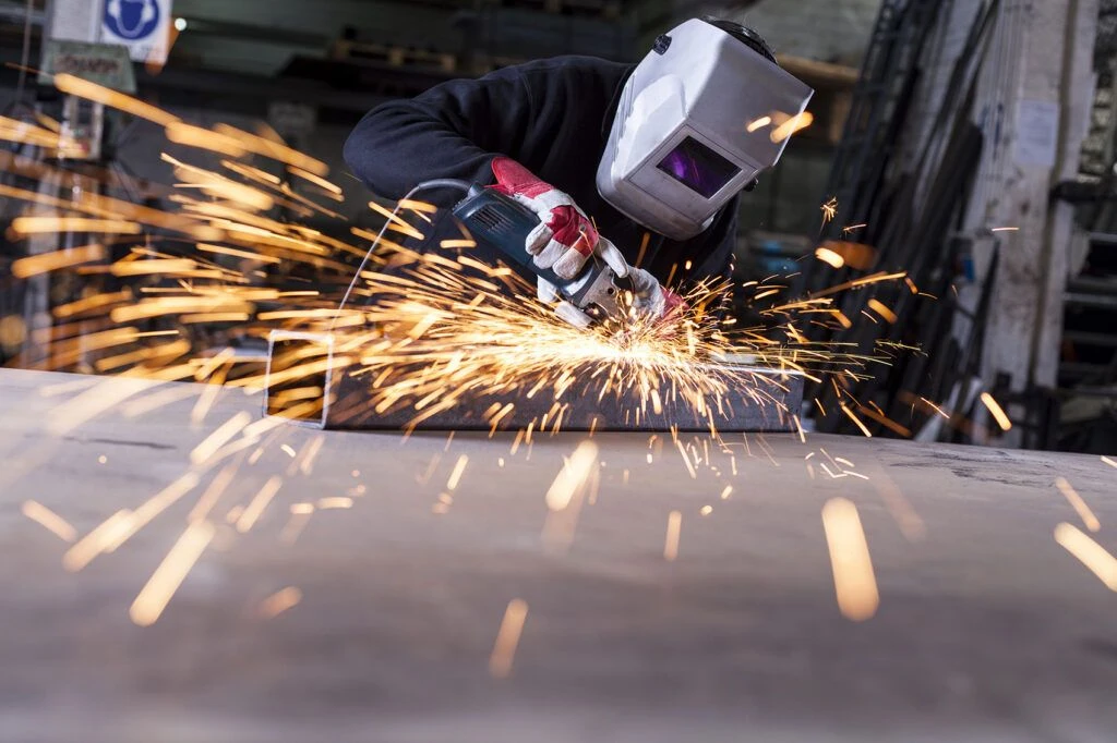 Person mit Schutzhelm und Arbeitshandschuhen bearbeitet ein Metallstück mit einem Winkelschleifer, während zahlreiche Funken sprühen.