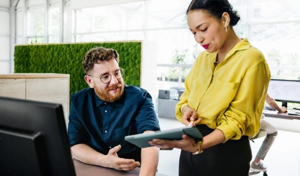 Ein Mann mit Brille und Bart sitzt an einem Schreibtisch und schaut auf ein Tablet, das ihm eine Frau in einer gelben Bluse zeigt. Im Hintergrund eine grüne Mooswand in einem hellen Büro.