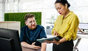 Ein Mann mit Brille und Bart sitzt an einem Schreibtisch und schaut auf ein Tablet, das ihm eine Frau in einer gelben Bluse zeigt. Im Hintergrund eine grüne Mooswand in einem hellen Büro.